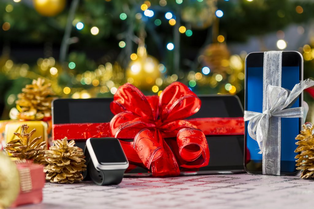Tablet-PC, Smartphone und Smartwatch mit Geschenken und Dekorationen vor einem Weihnachtsbaum.